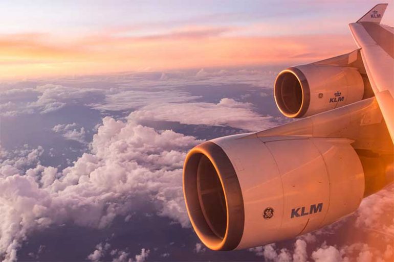 Photo is taken from an aeroplane window. It shows the clouds and sky with a beautiful orange/red tint of sunset and the plan engines.