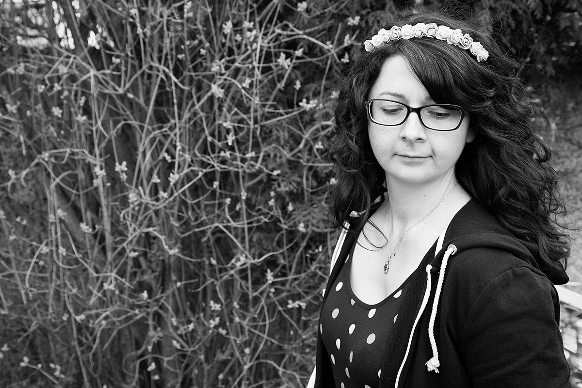 A black and white photograph of Donna in the right side of the frame. She has shoulder length curly hair accessorised with a flower crown. She wears glasses and is wearing a polka dot dress and open zipped hoodie. She is looking down.
