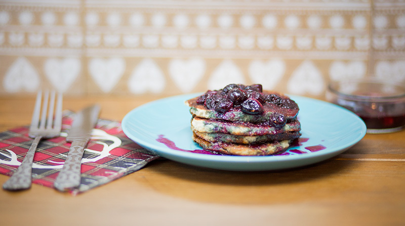 Photograph shows dairy and gluten free protein pancakes with a blueberry topping on a blue plate with a napkin and fork and knife at the side.