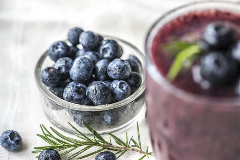 Photo shows a small bowl filled with blueberries in focus and a glass with a blueberry smoothie out of focus.