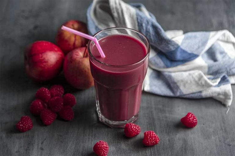 Photo shows a clear glass containing a red berry smoothie with a pink straw.. In the background are apples and raspberries and a dish cloth.