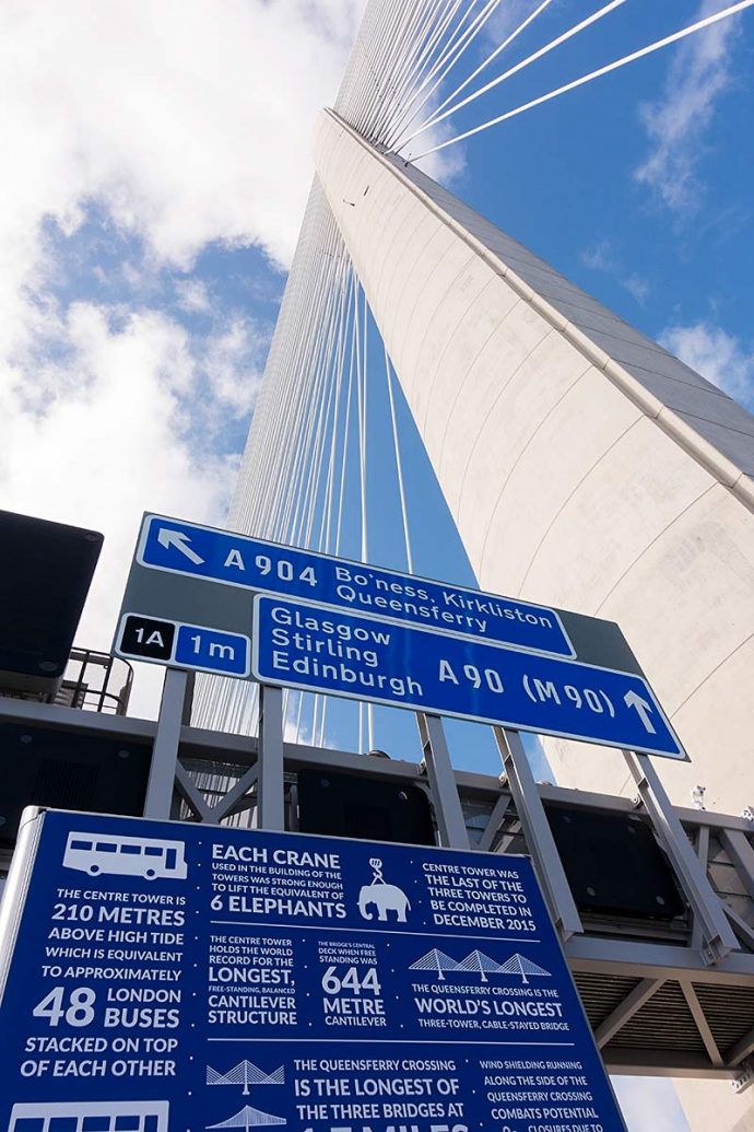 Photograph shows the road signage plus an informative sign with facts about the construction of the Queensferry Crossing bridge with the support tower and cables behind it.