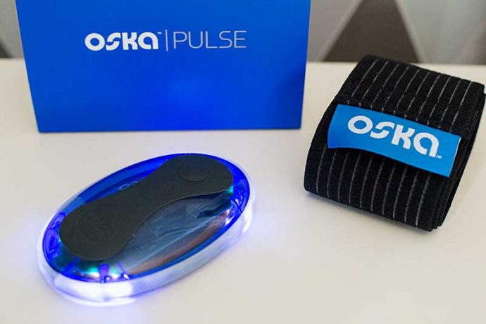 Photograph shows the Oska pulse pain relief device. It is a palm sized, oval shaped gadget that is blue and black and lights up when in use. Also featured is the Oska Pulse band (for wearing the device). The Oska Pulse logo is displayed behind on a blue box.