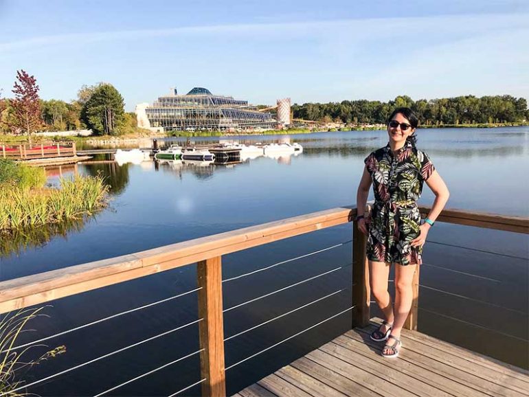 Photo shows Donna standing on a dock by the edge of a lake resting against a wooden fence. She is wearing a leaf print playsuit, sunglasses and her dark hair is tied back in a ponytail. In the distance behind her there is the glass Aqualagon building at Villages Nature, Paris which houses swimming pools and water slides.