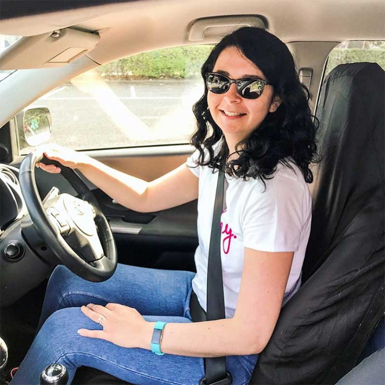 Photo shows Donna sitting in the driving seat of a car with one hand on the wheel and one resting on her leg. Donna has shoulder-length dark curly hair and is wearing sunglasses, a white t-shirt with the slogan "hey" and blue jeggings.