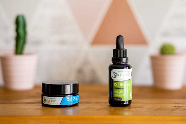 Photo shows the Nganic 1000mg CBD oil tincture and salve on a wooden shelf. There are are two cacti in the background in pink pots and marble, copper, grey and pink triangle patterned wallpaper.