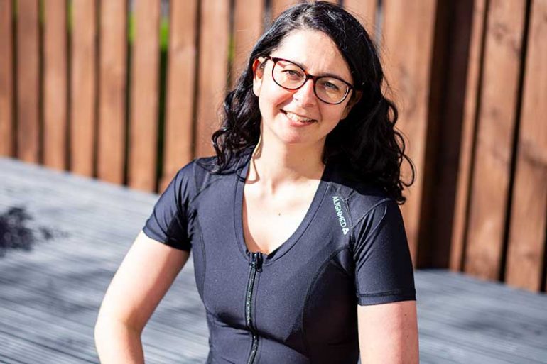 Photo shows Donna wearing the black Active Posture 2.0 shirt with front zip. Donna is looking and smiling at the camera and has long, dark curly hair and wears glasses. The photo is cropped at her waist.