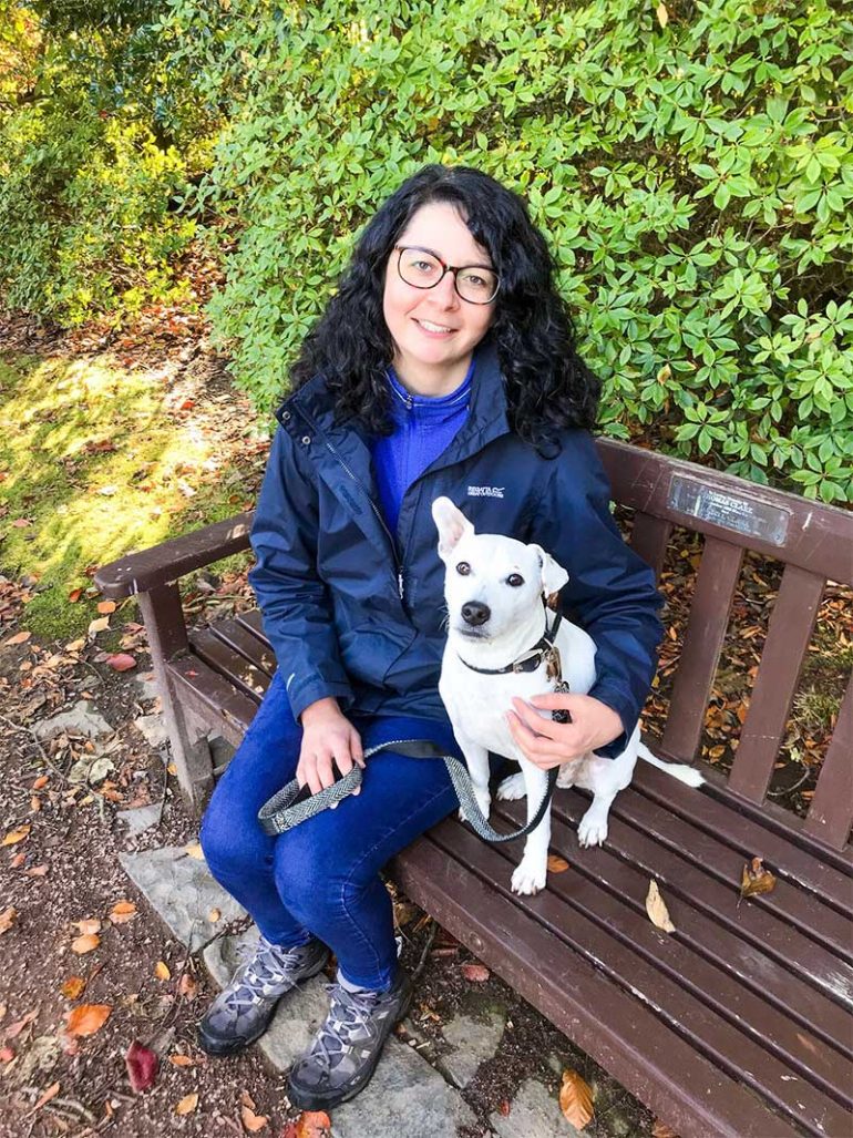 Photo shows Donna, a woman with shoulder-length dark curly hair, sitting on a bench with her dog Oscar, a white Jack Russell Terrier cross sat net to her. She has her arm around him and they are both looking at the camera.