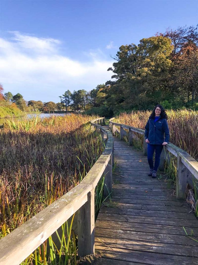 Photo shows Donna on a wooden boardwalk on a nature walk. She is leaning against a wooden railing and looking at the camera. In the background are trees and a beautiful blue sky.