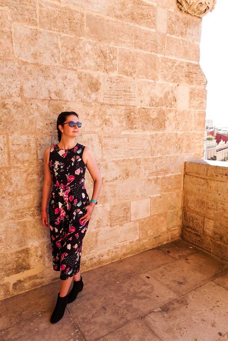 Photo shows Donna stood against a brick wall in the Serrano Towers in Valencia. She wears a black and floral jumpsuit, black ankle boots and dark sunglasses. She has her hair in dutch braid pigtails and is looking off to the right.