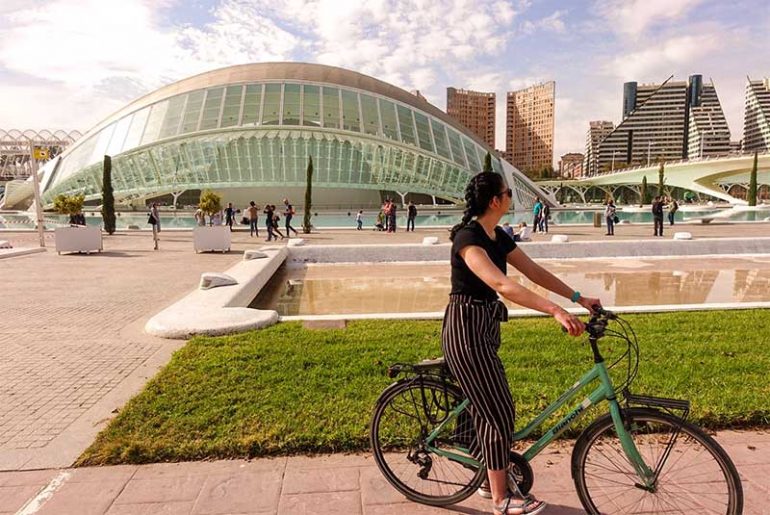 Photo shows Donna sat on a teal bike looking to the right away from the camera. Behind her is a concrete pool of water (water feature) and a building at the City of Arts and Sciences in Valencia that has been designed to resemble a human eye.