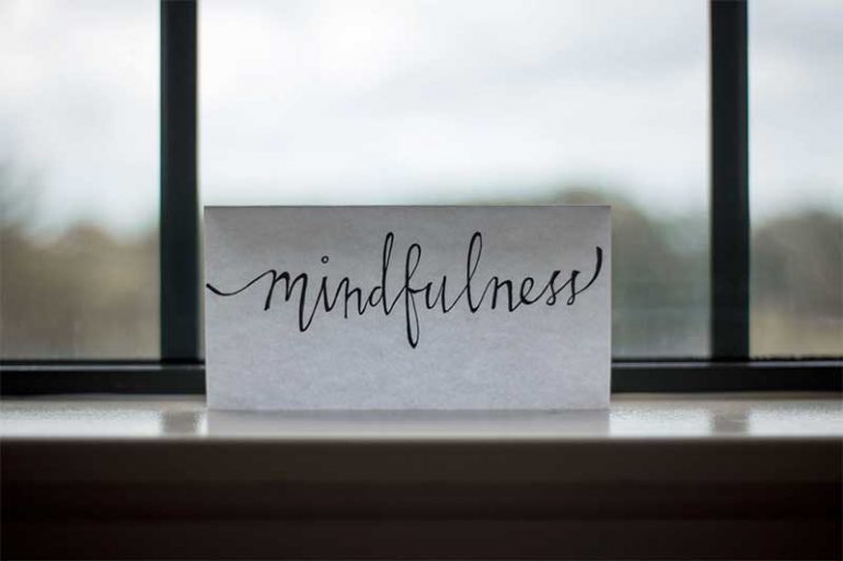 Photo shows a white piece of paper resting on a windowsill with the word "mindfullness" written in a calligraphy style with black ink. There is a window behind the note but the view out of it is blurred.