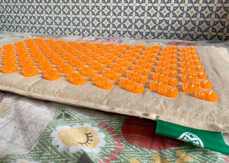 Photo shows a Mindfulness Mat: a linen cushioned mat with orange spiky triangular massagers covering the surface. It is placed on a bed with an owl print duvet and there is a blue/white floral patterned wallpaper behind.
