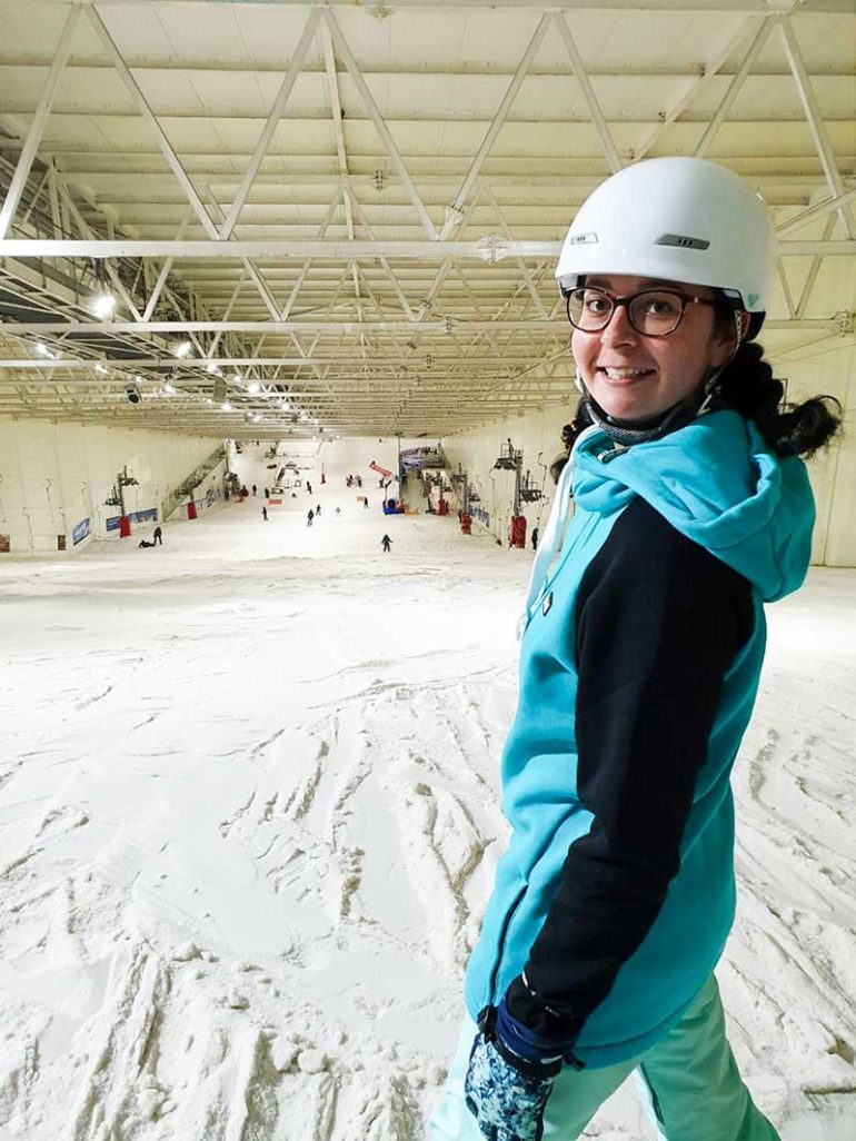 Photo shows Donna stood at the top of the indoor snow slope. She has turned her body towards the camera and is smiling. The photograph is cropped at her knee so you cannot see the snowboard attached to her feet. Donna wears a teal hoodie with black sleeves and a white helmet. Her dark hair is braided.