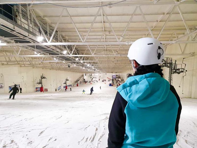 Photo is taken from behind Donna at the top of the indoor snow slope. The photo is cropped at her waist and Donna is looking ahead, down the slope. Donna wears a teal hoodie with black sleeves and a white helmet.
