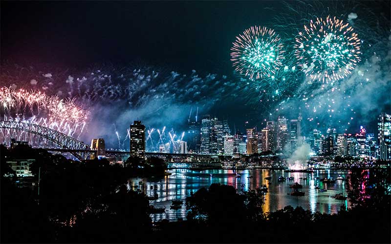 Photograph shows fireworks over a river with the silhouette of a bridge and city in the background.