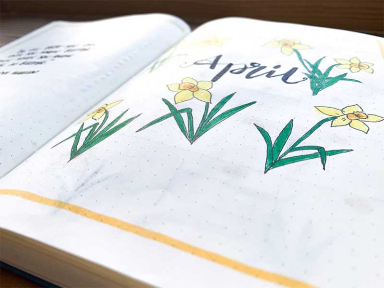 Shallow focus photograph taken across my bullet journal cover page showing the word April written in calligraphy surrounded by illustrative daffodils and a yellow border.
