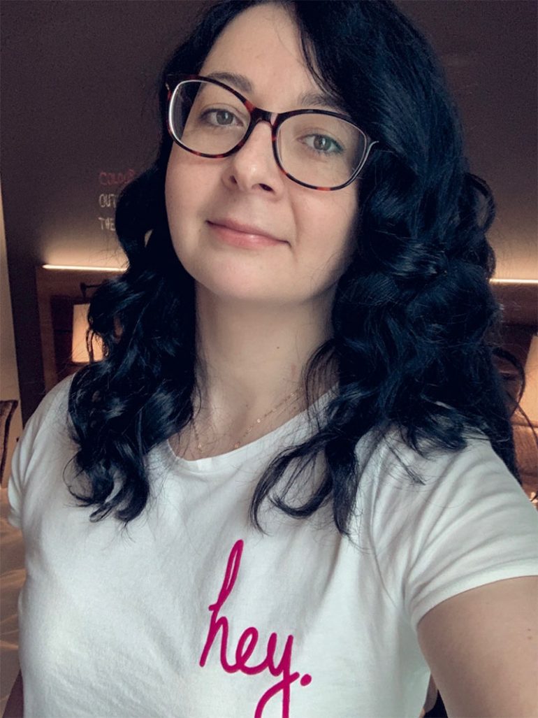 Selfie of Donna, a white woman with shoulder-length dark wavy hair. She wears glasses and is wearing a white t-shirt with the slogan "hey" in pink.