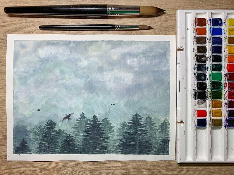 Photo shows a watercolour painting of a bird's eye view of a misty forest with three birds flying in the sky. Sat next to the painting are watercolour pans and two watercolour brushes