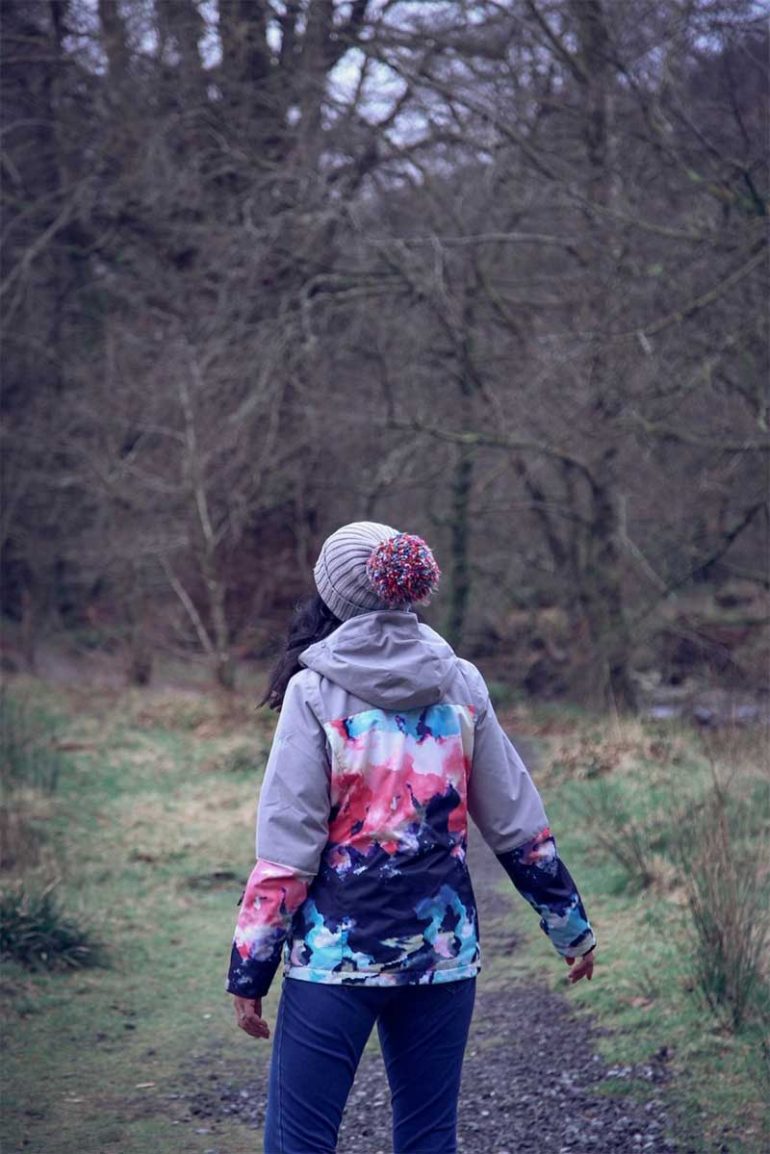 Photo of Donna taken as she is walking away from the camera along a path in the countryside. She is wearing a grey beanie with large multi-coloured pom pom and a colourful jacket with a watercolour pattern.