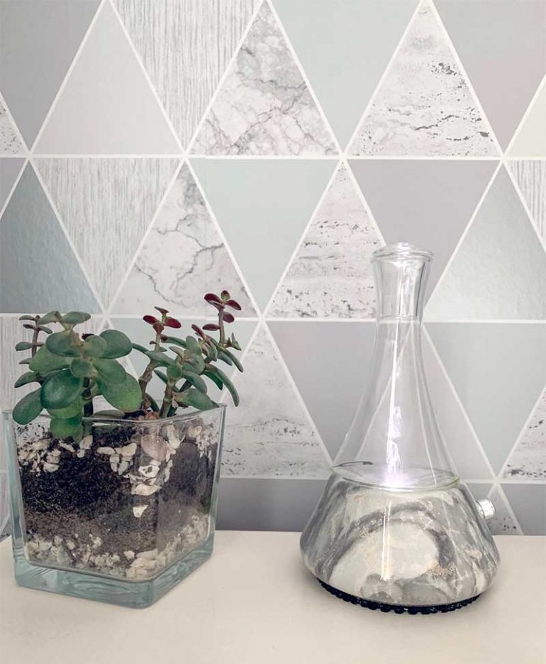 Photo shows the opulence grey marble nebuilizing diffuser sat on a bedside table next to a succulent plant in a glass cube. This is the diffuser I chose as a beginner to essential oils.
