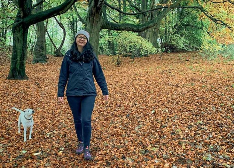 Donna walks down a path covered in fallen leaves with beautiful autumnal colours. Behind her, looking up at her, is her little white dog Oscar.