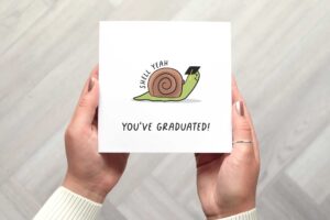 This card features a cute illustrated snail who is smiling and wearing a graduation cap. The words shell yeah follow the curve of its shell and youve graduated is written beneath the illustration.