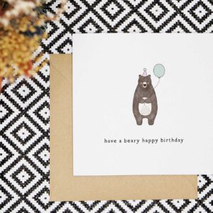 This birthday card features a hand illustrated brown bear wearing a lilac party hat with a matching balloon in its mouth. It reads have a beary happy birthday.