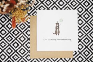A fun birthday card featuring a cute otter that is wearing a green party hat with grey pom pom that is holding a green balloon. Card reads have an otterly awesome birthday.
