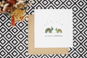 This cute pun card features two illustrated tortoises who are facing each other and wearing party hats. Above them are grey musical notes and beneath them are the words its time to shellebrate