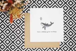 This birthday card features a hand illustrated humpback whale wearing a green party hat with a matching balloon in its mouth. It reads have a Whaley great birthday.