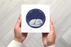 This card features a circular illustration in the centre of an arctic fox sleeping in the grass with a background of a starry nights sky and florals around each side of the circle.