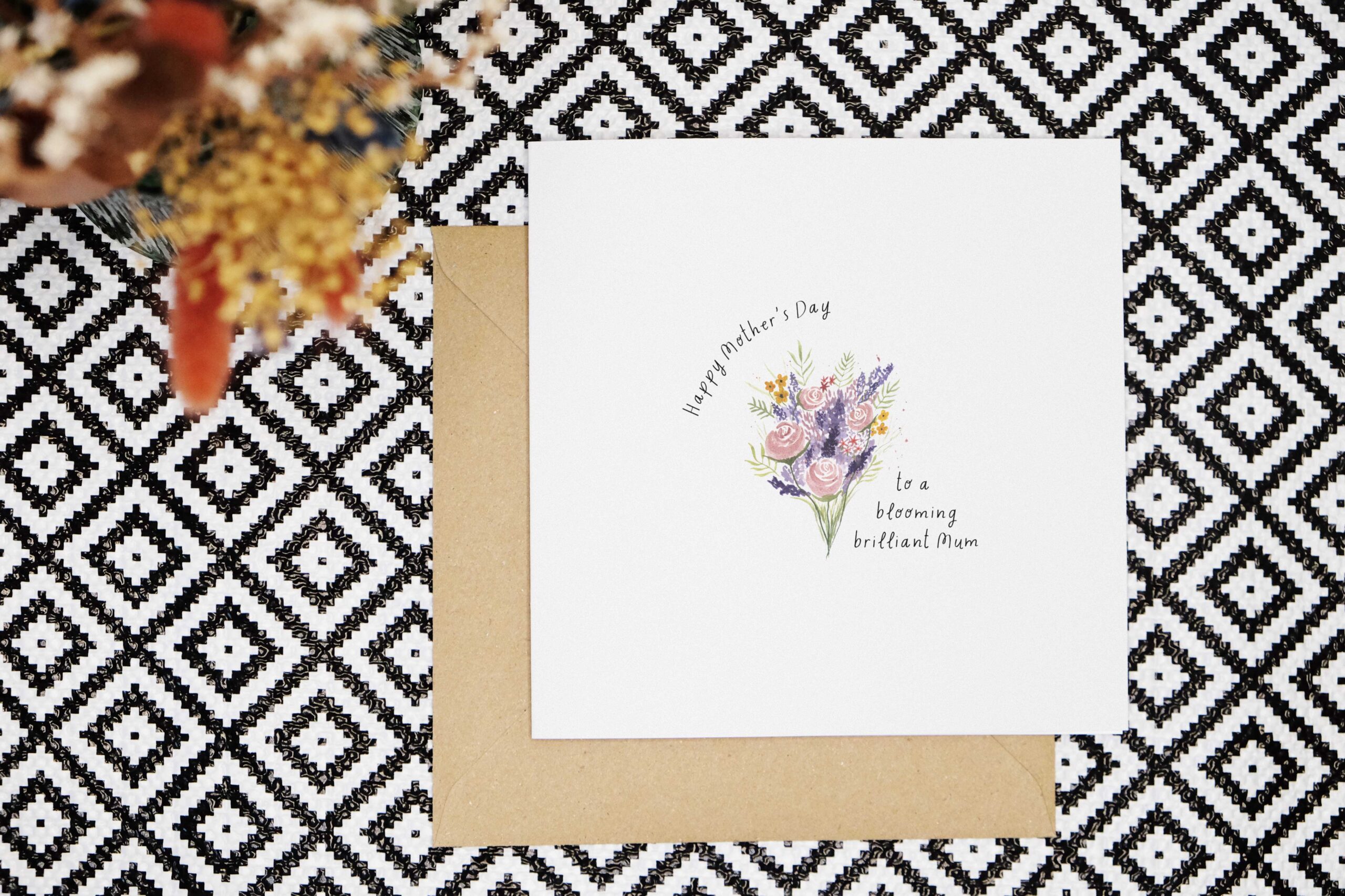 This card features a watercolour bouquet painted in a loose style with pink, purple, red and yellow flowers and green leaves. The card reads Happy Mothers Day to a blooming brilliant Mum in a black handwritten font