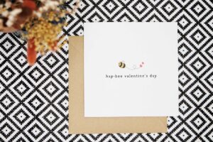 A cute valentines card featuring an illustrated bee leaving a trail behind it with two love hearts at the end. The card reads hap-bee valentines day