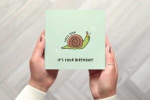 This card features a cute illustrated snail who is smiling and wearing a party hat. The words shell yeah follow the curve of its shell and its your birthday is written beneath the illustration. There are two white stars with four points on either side of the snail. The card is background is a bright pastel green.