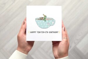 40th birthday card features a cute illustrated tortoise sitting in a teal tea cup with white polka dots. It wears a yellow party hat with 40 on it. There is a tor-tea label from a tea bag hanging on the side and the words happy tor tea eth birthday