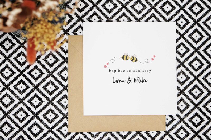 A cute anniversary card featuring two illustrated bees flying towards each other. Each bee has a trail of dashes behind them with two love hearts at the end. The card reads hap bee anniversary and is personalised with the couples names