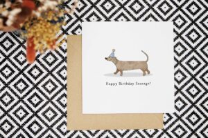 This cute and funny birthday card features an illustrated dachshund wearing a party hat who is strutting along. There is a shadow under the dog. Beneath this are the words happy birthday sausage