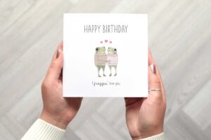 Cute birthday card that features two frogs wearing knitted jumpers who are looking at each other and holding hands. Three love hearts are above their heads. Card reads happy birthday I froggin love you.