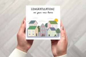 Card shows a little illustrated scene with 5 houses, 3 in front and 2 behind. There are trees and shrubs dotted throughout the illustration. The sun can be seen peeking up behind one of the roofs and clouds are in the background. A cute illustration.