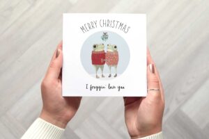 Cute Christmas card that features two frogs wearing knitted jumpers who are looking at each other and holding hands while standing under mistletoe. There is a circular light blue background. Card reads Merry Christmas I froggin love you.