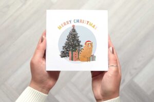 Christmas card that features a Christmas tree with multi coloured lights, a ginger cat wearing a Santa hat and two Christmas presents. There is a circular light blue background. Card reads Merry Christmas in a bold colourful font.