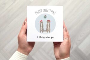 Cute Christmas card that features two otters wearing Santa hats who are standing under mistletoe. There is a circular light blue background. Card reads Merry Christmas I utterly adore you.