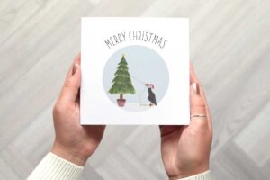 Cute Christmas card that features a puffin wearing a Santa hat that is putting lights on a Christmas tree. There is a circular light blue background. Card reads Merry Christmas.
