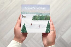 The card shows a wintery, illustrated scene with two frogs called the froggos ice skating on a frozen lake framed by two trees. In the middle ground is a forest and a wooden cabin with snowman outside. In the background is a beautiful sunset.