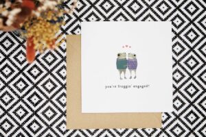 Engagement card features two frogs wearing knitted jumpers who are looking at each other and holding hands while standing under 3 love hearts. One has an engagement ring on its hand. Card reads youre froggin engaged!