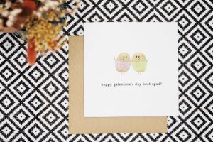 This cute pun card features two potatoes dressed in knitted jumpers, one pink with love hearts and one green, stood with one arm around each other and one arm in the air. The card reads happy galentines day best spud!