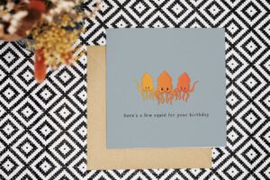 A funny birthday card with blue grey background and three cute illustrated squid in the middle that are yellow, orange and red in colour. The card reads heres a few squid for your birthday