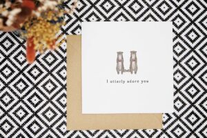 Cute Valentines card that features two otters holding love hearts standing next to each other with their tails resting together. Card reads I otterly adore you.