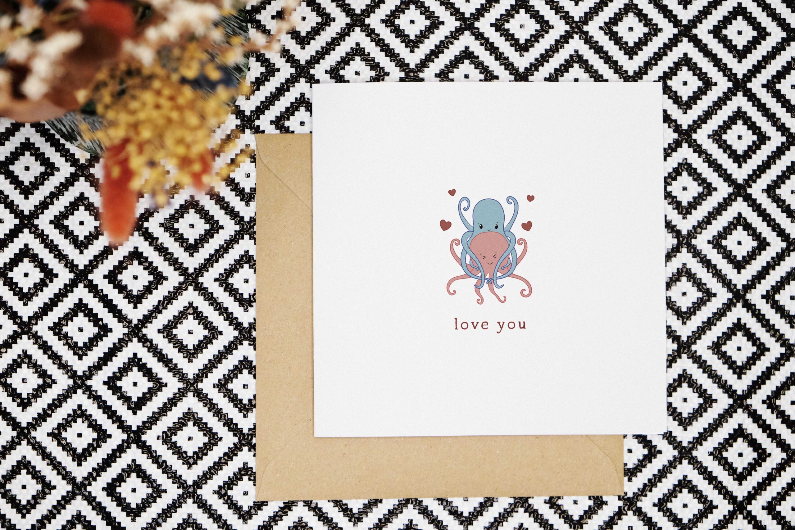 A cute love you card featuring a hand illustrated pink octopus that is being hugged from behind by a blue octopus. There are four love hearts around them and the word love you in a red font below the illustration.
